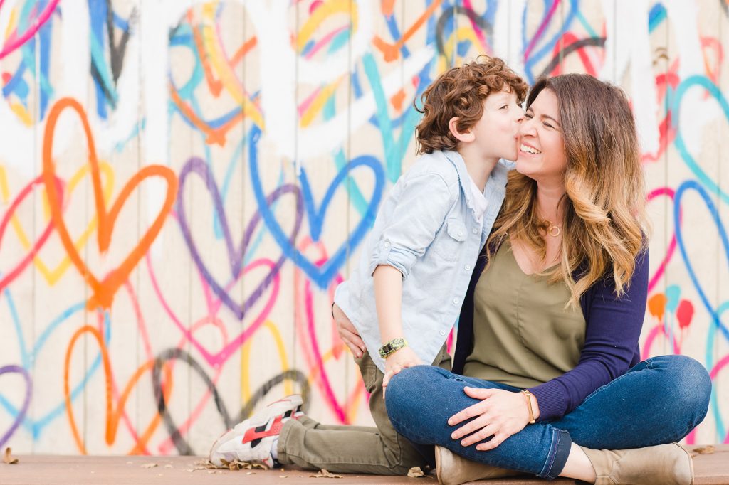 Grant gives mom a kiss in front of a rainbow heart wall.